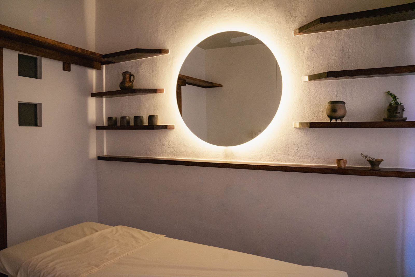 A serene bedroom at Casa de Portales with a round illuminated mirror and wooden shelves.